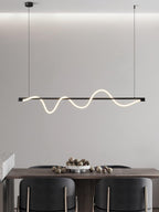 Black Clérachic linear pendant with a glowing wave, hanging above a walnut table set with a bowl of nuts and dark chairs.