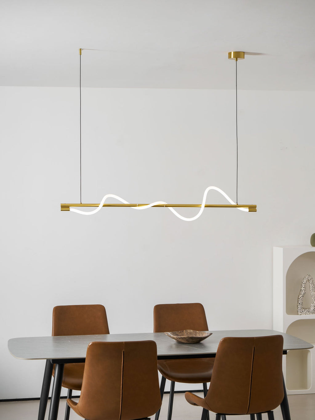 A brass Clérachic linear pendant with white silicone waves, hung over a grey table and rust leather chairs.