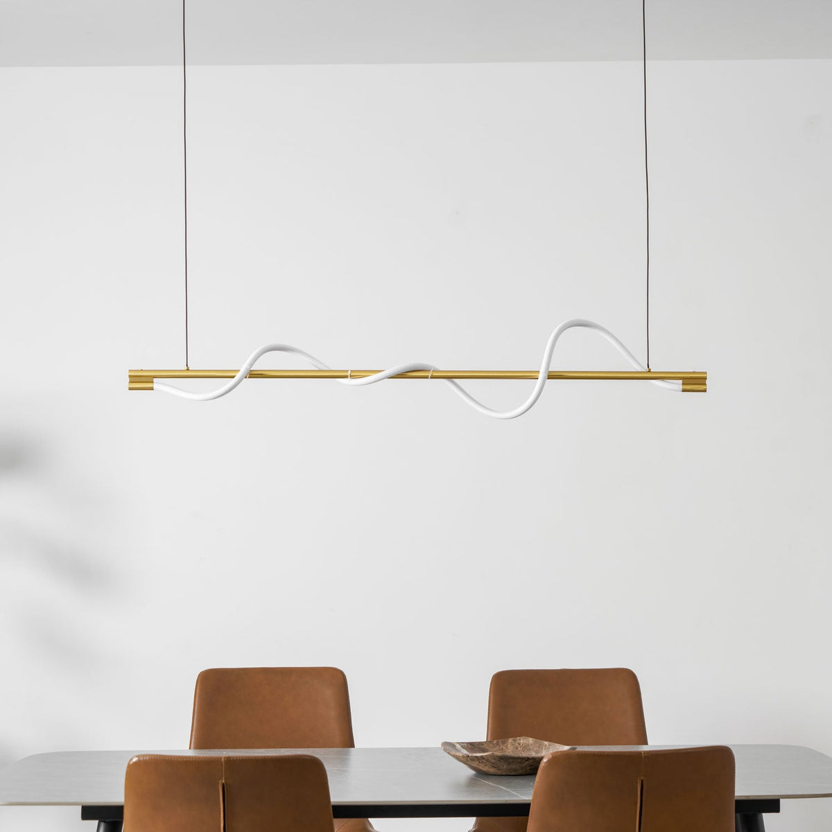 Brass Clérachic linear pendant with a white silicone wave, in front of a white wall and a table with rust chairs.