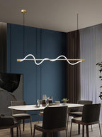 Clérachic brass linear pendant lit above a marble table and dark chairs, in a dining room with deep blue walls.