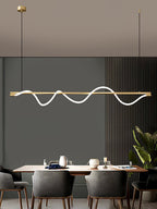 Brass Clérachic linear pendant with a lit wave, above a wood table in a dining room with grey slatted walls.