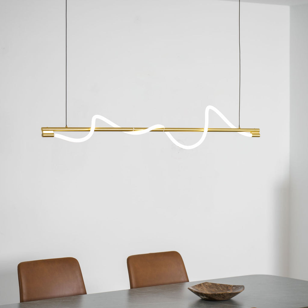 Clérachic brass linear pendant with glowing waves, above a grey table, tan chairs and a wooden bowl.