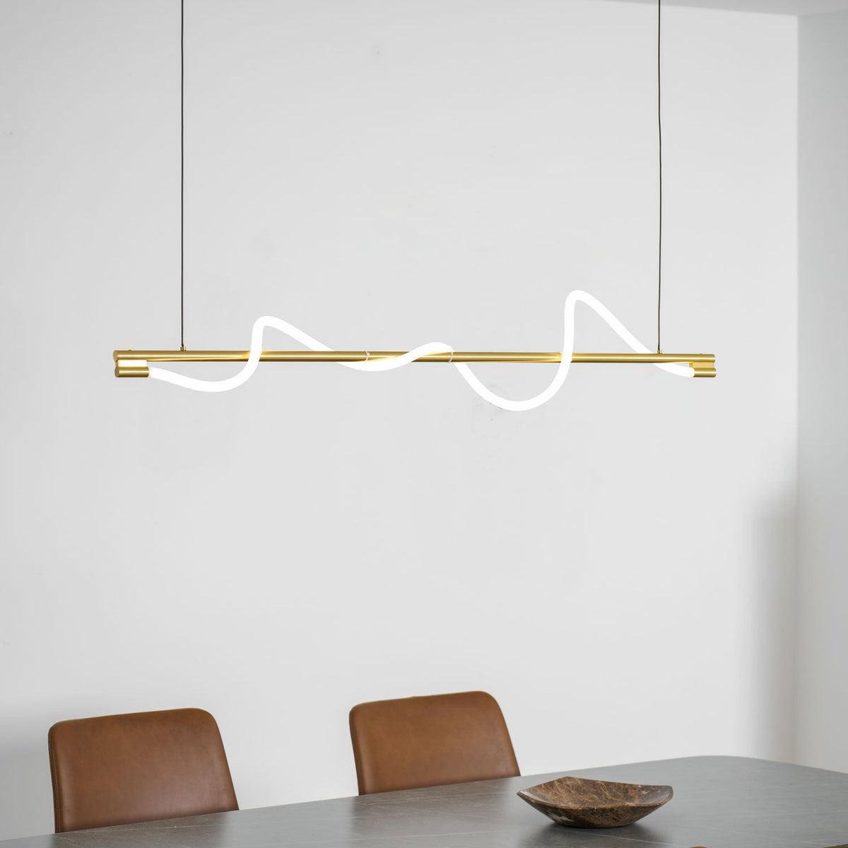 Clérachic brass linear pendant with glowing waves, above a grey table, tan chairs and a wooden bowl.