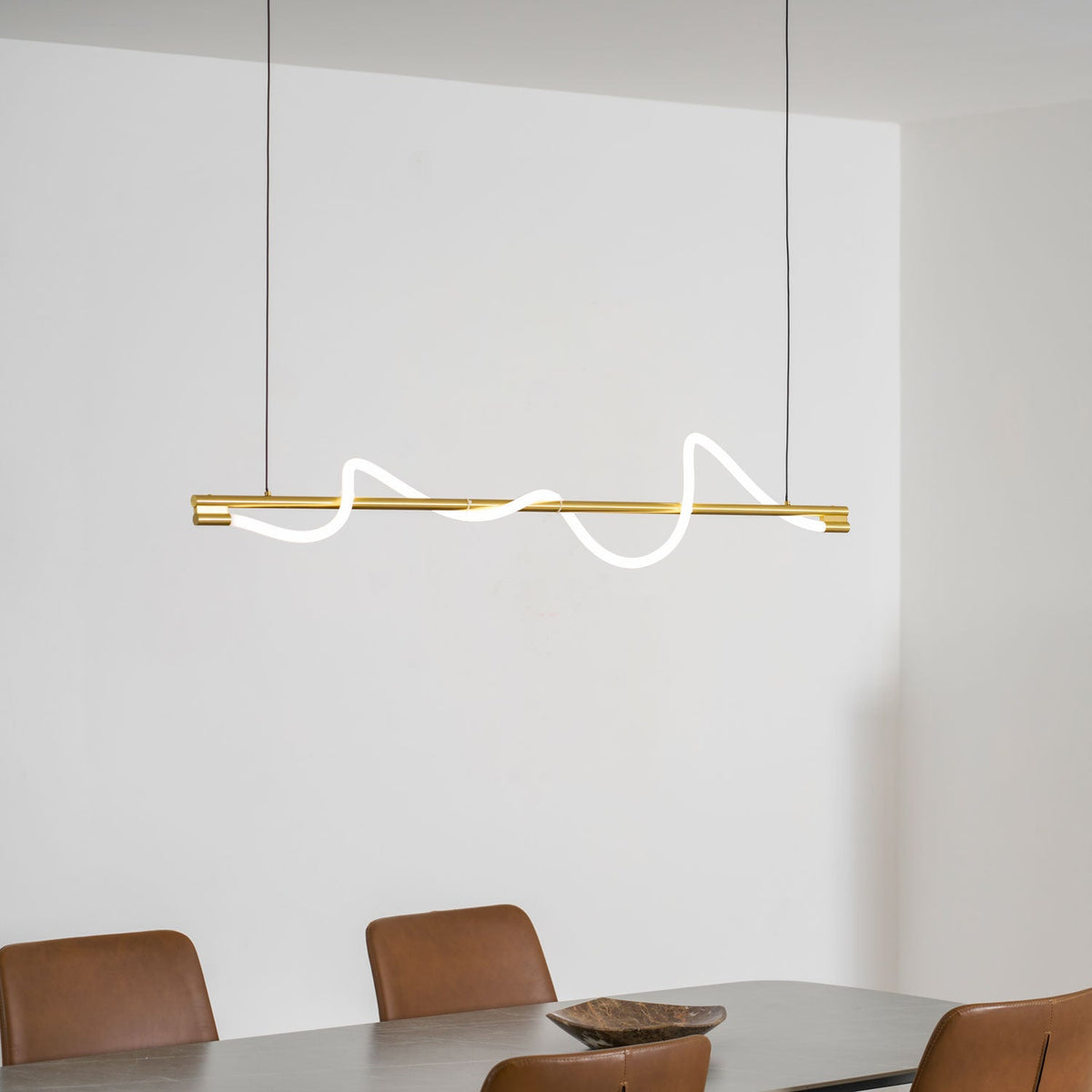 Brass Clérachic linear pendant with a glowing wave, over a table with tan chairs, in a white room.