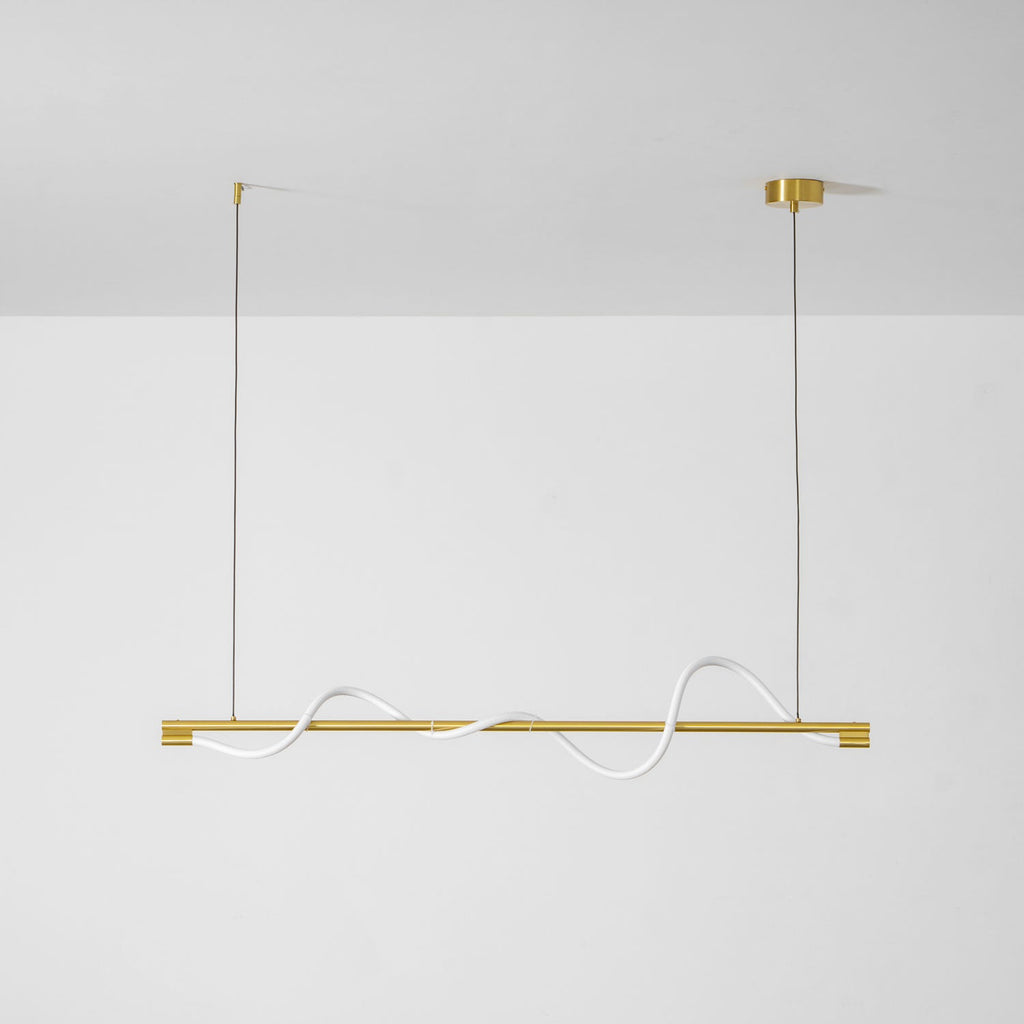 Unlit brass Clérachic linear pendant with a white silicone wave, on a white wall with brass canopies.