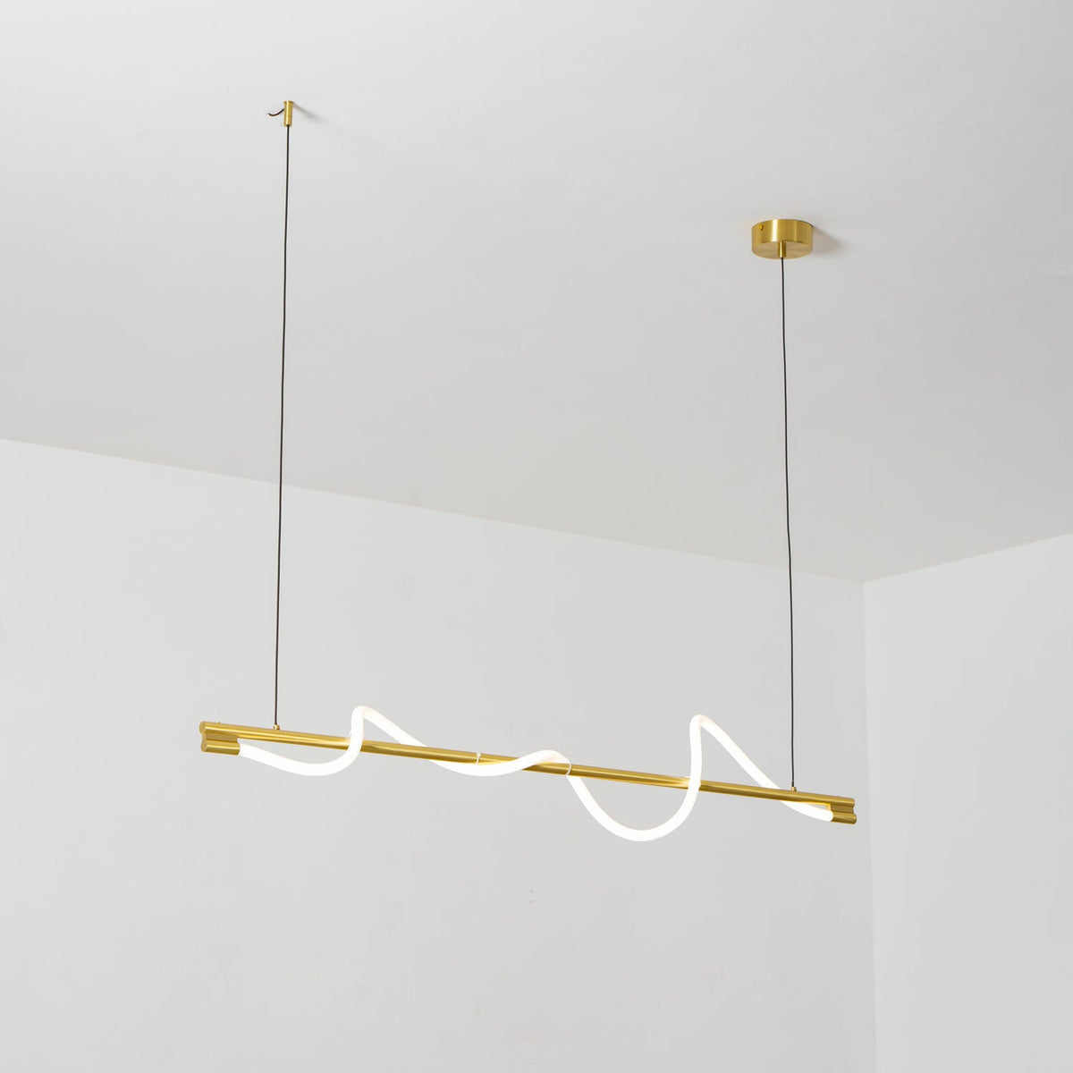 Looking up at a brass Clérachic linear pendant with a glowing wave, hung from a white ceiling.