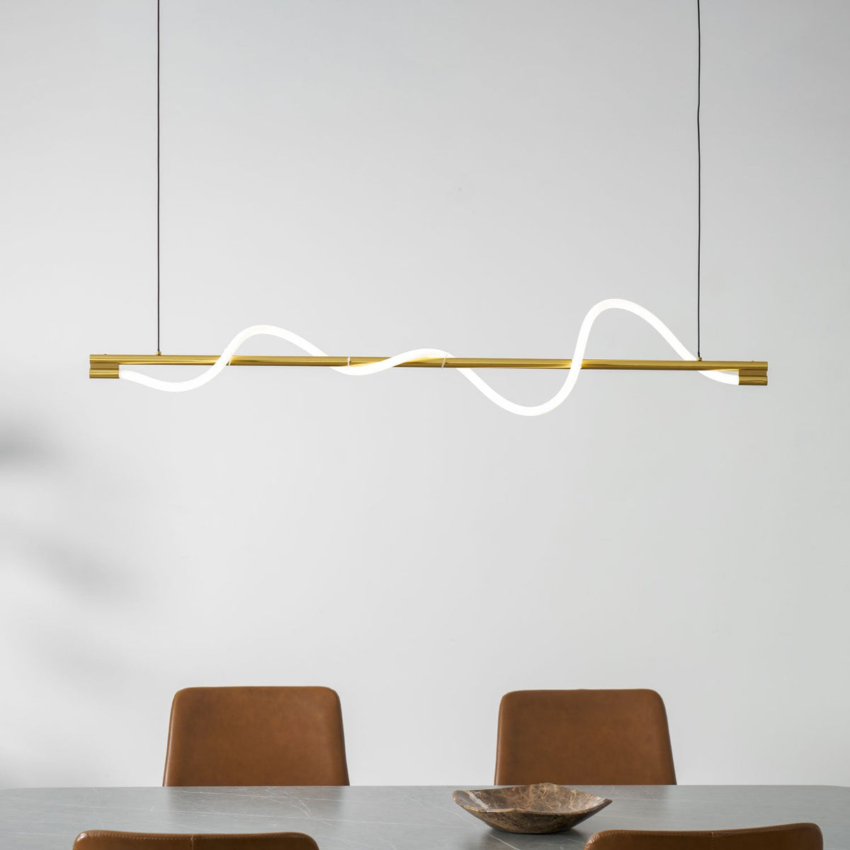 Brass Clérachic linear pendant with a lit wave, above two rust leather chairs and a wooden bowl.