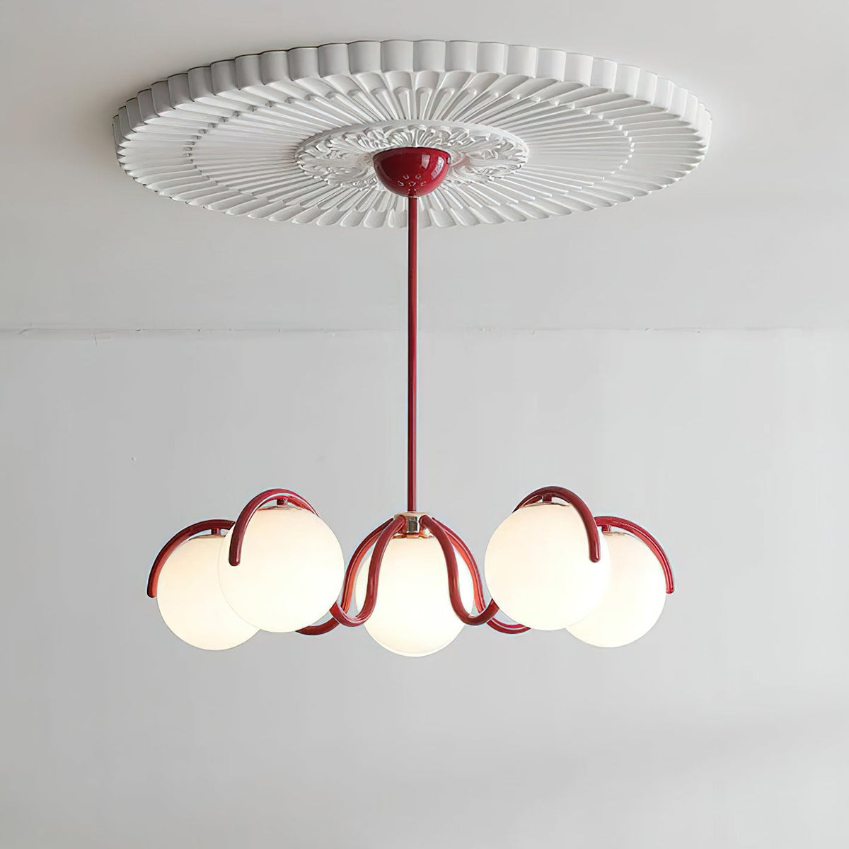 A red Clérachic chandelier with six glass globes hung from a white pleated ceiling rose against a pale wall.