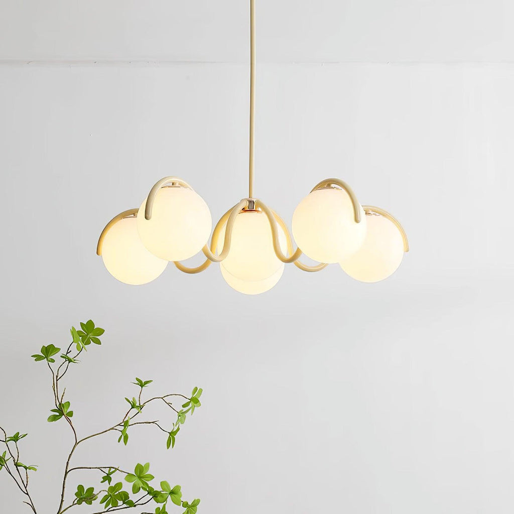 Clérachic chandelier in creamy yellow, with six lit globes on wavy arms and a green sprig in the lower corner.