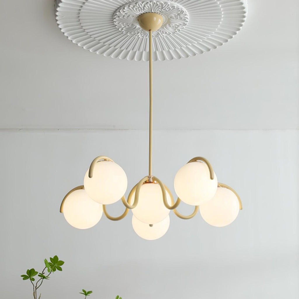 A Clérachic chandelier in creamy yellow, hung from a ceiling rose with its globes lit and a sprig of leaves at the corner.