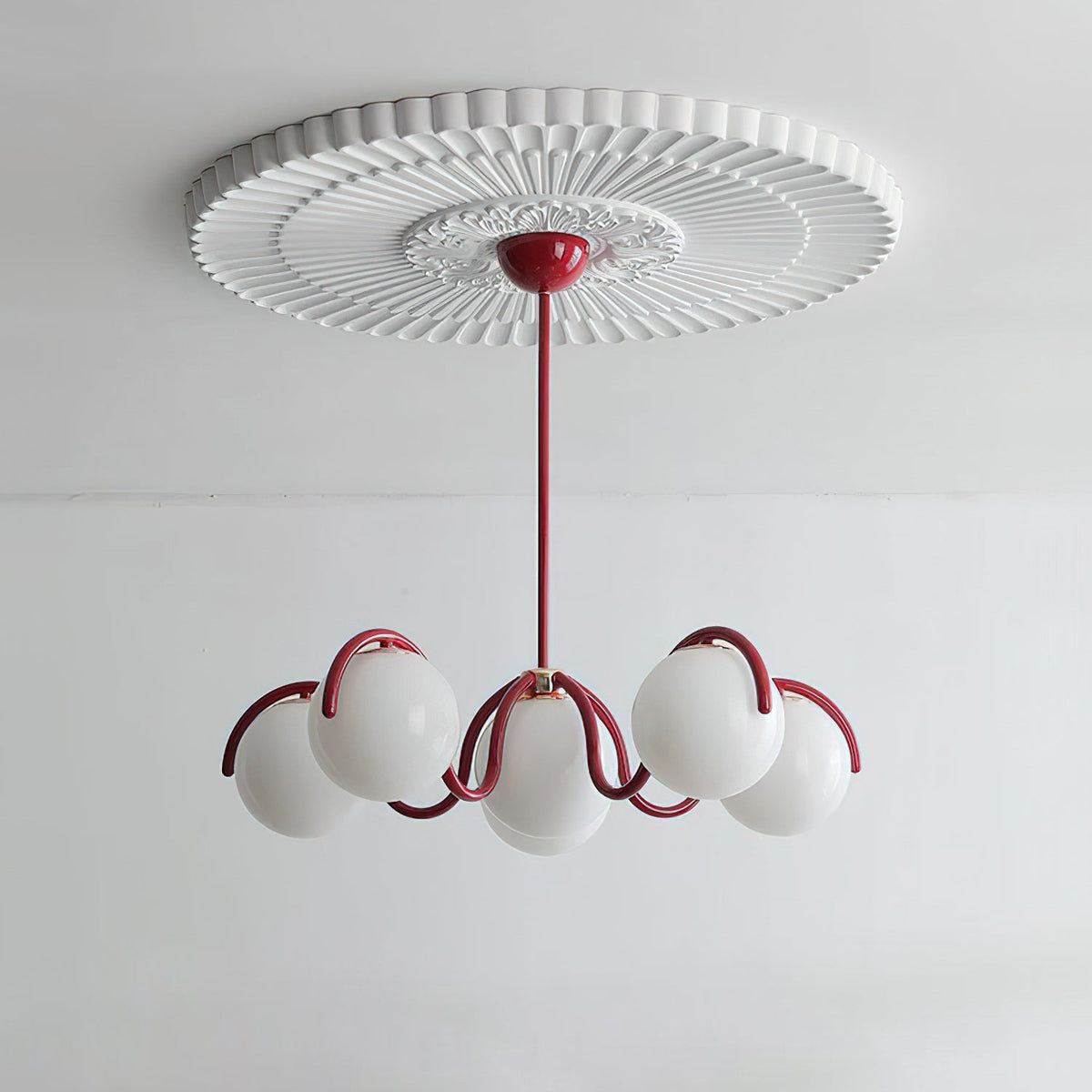 Red Clérachic chandelier with its globes unlit, hung under a pleated ceiling rose against a pale wall.