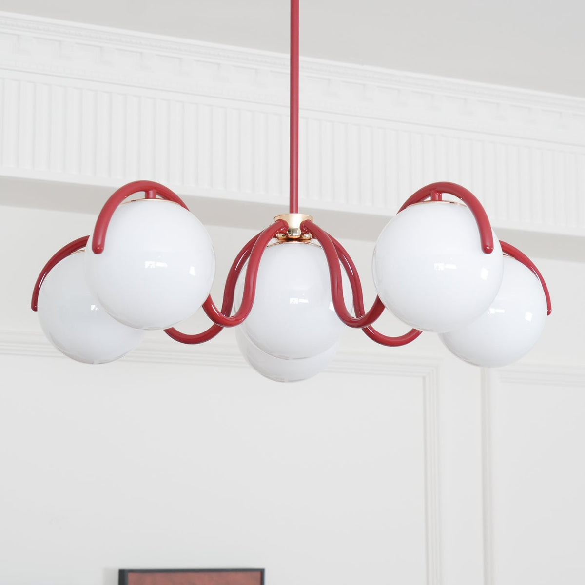 A red Clérachic chandelier seen from below against a white panelled wall, with a coved ceiling edge above.