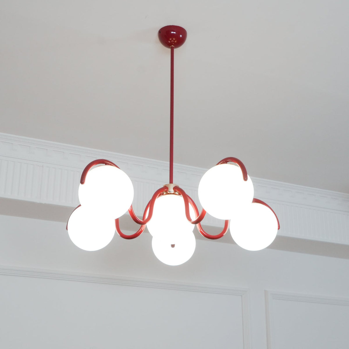 A red Clérachic chandelier from below with a round red canopy, its globes lit warm against a white cornice.