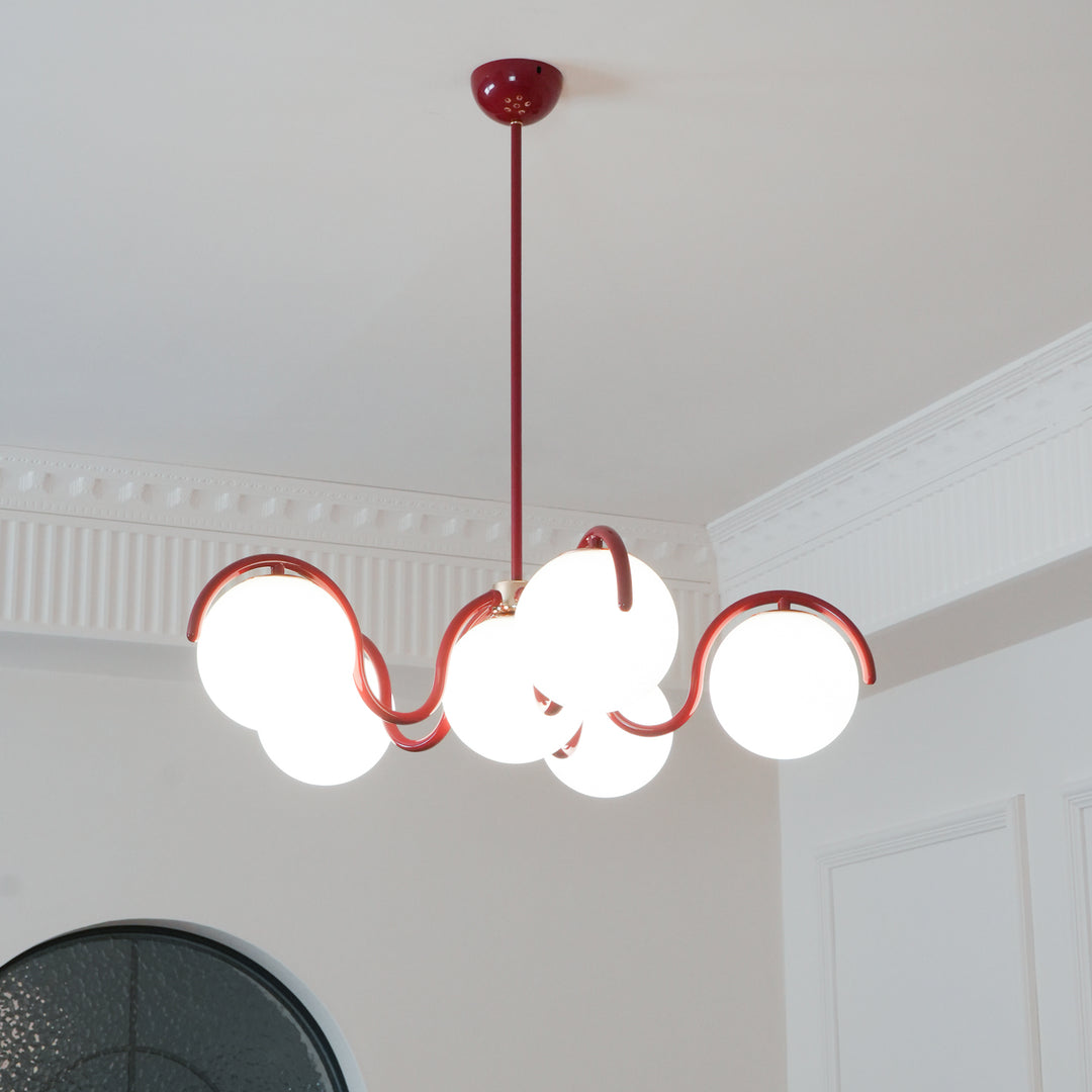 A lit red Clérachic chandelier seen from below, with its curving arms and globes in front of a white panelled wall.