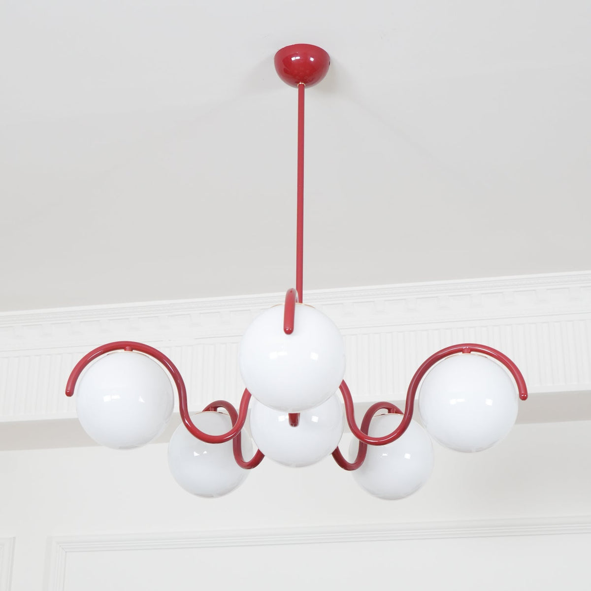 A red Clérachic chandelier with unlit white globes, seen from below against a white cornice and pale wall.