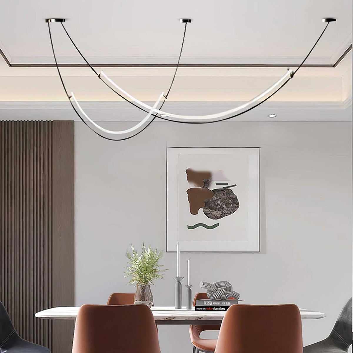 Clérachic pendant with two glowing LED curves above a marble-top table and rust-coloured chairs, a framed abstract print behind.