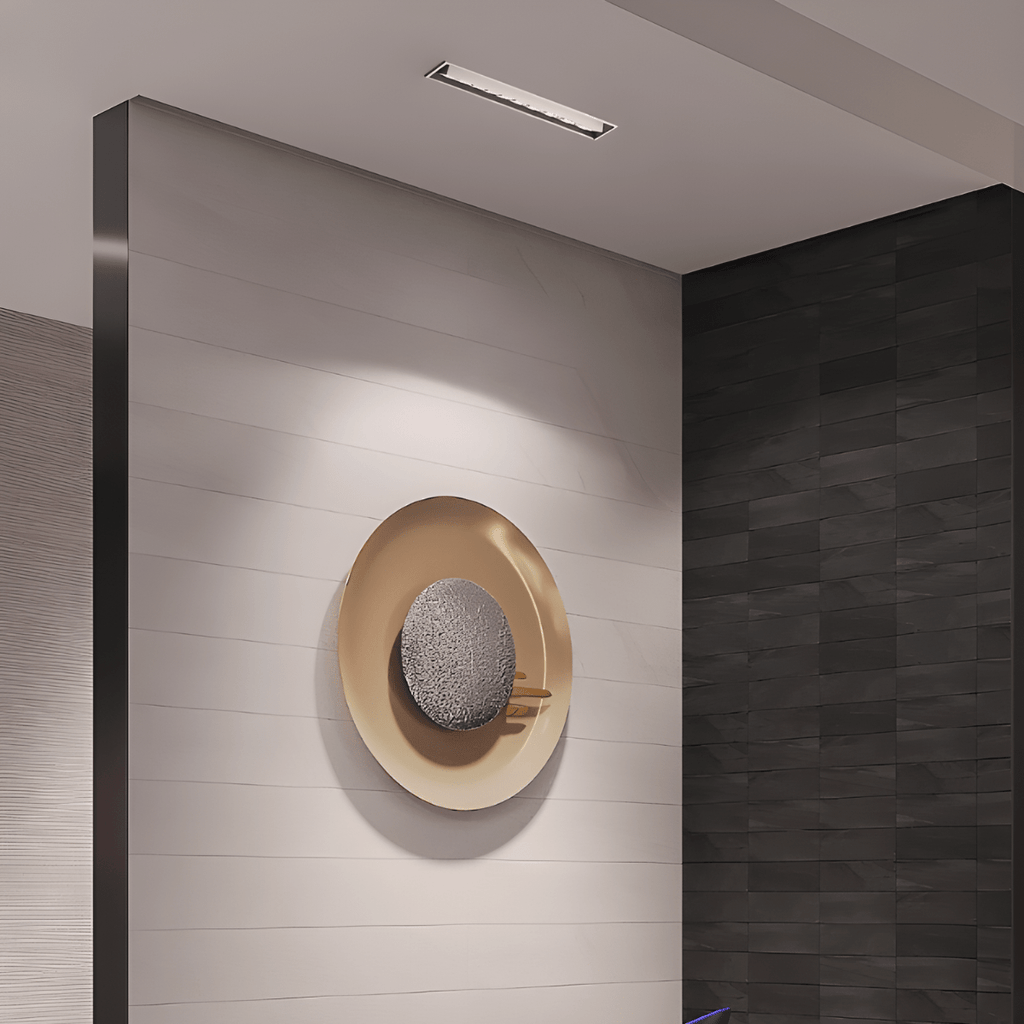 Clérachic slim white recessed downlight in a ceiling above a wall with a round brass-and-stone art piece beside a dark tiled corner.