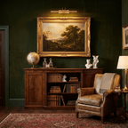 Above a gold-framed landscape painting on a dark green wall, this gold Clérachic wall light shines down across the canvas.