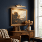 Gold Clérachic wall light above a gold-framed landscape on a navy wall, with a wooden desk, dried flowers and books below.