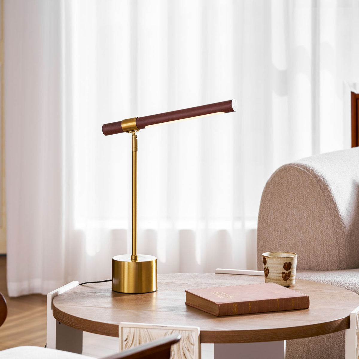 Clérachic table lamp on a round wooden side table beside a book and a small cup, with sheer white curtains behind.