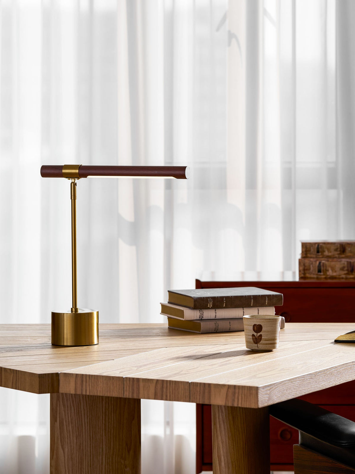 Low-angle view of this Clérachic desk lamp on a pale oak table, its walnut bar level above a stack of books and a cup.