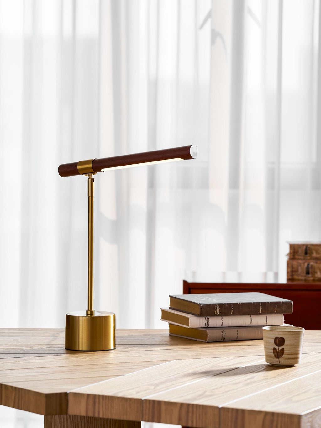 Upright shot of this Clérachic lamp on a herringbone wooden desk, with books and a patterned cup to its right.