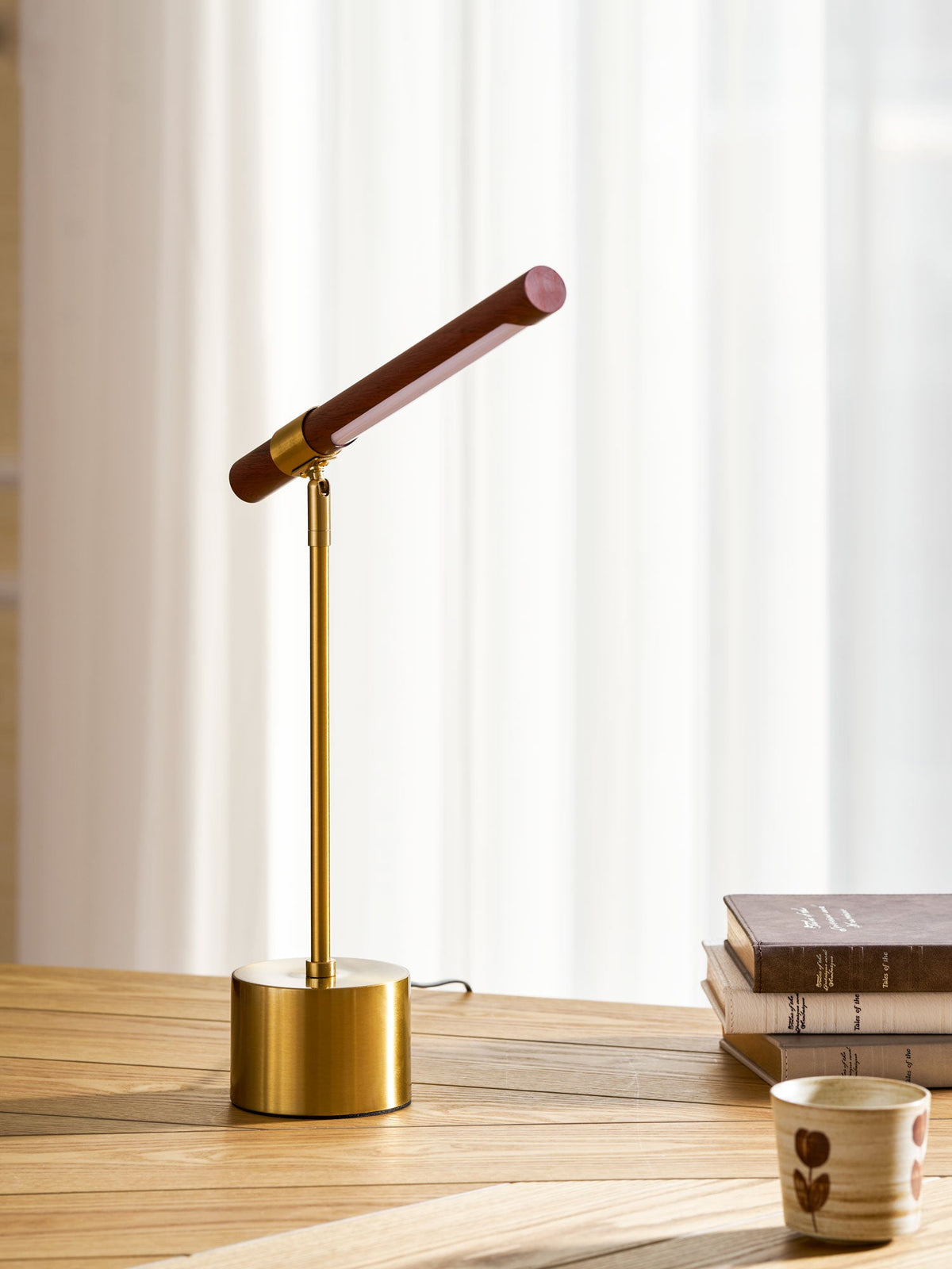 This Clérachic lamp with its walnut bar angled steeply upward, on a wooden desk in front of white curtains.