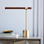 Clérachic table lamp on a grey cabinet by a pale wall, its walnut bar level above a brass base, with books and a brass dish.