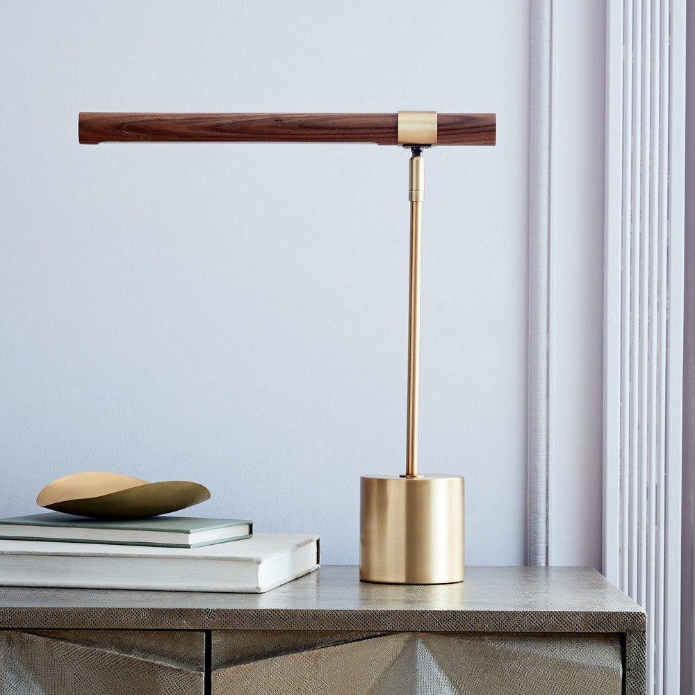 Clérachic table lamp on a grey cabinet by a pale wall, its walnut bar level above a brass base, with books and a brass dish.