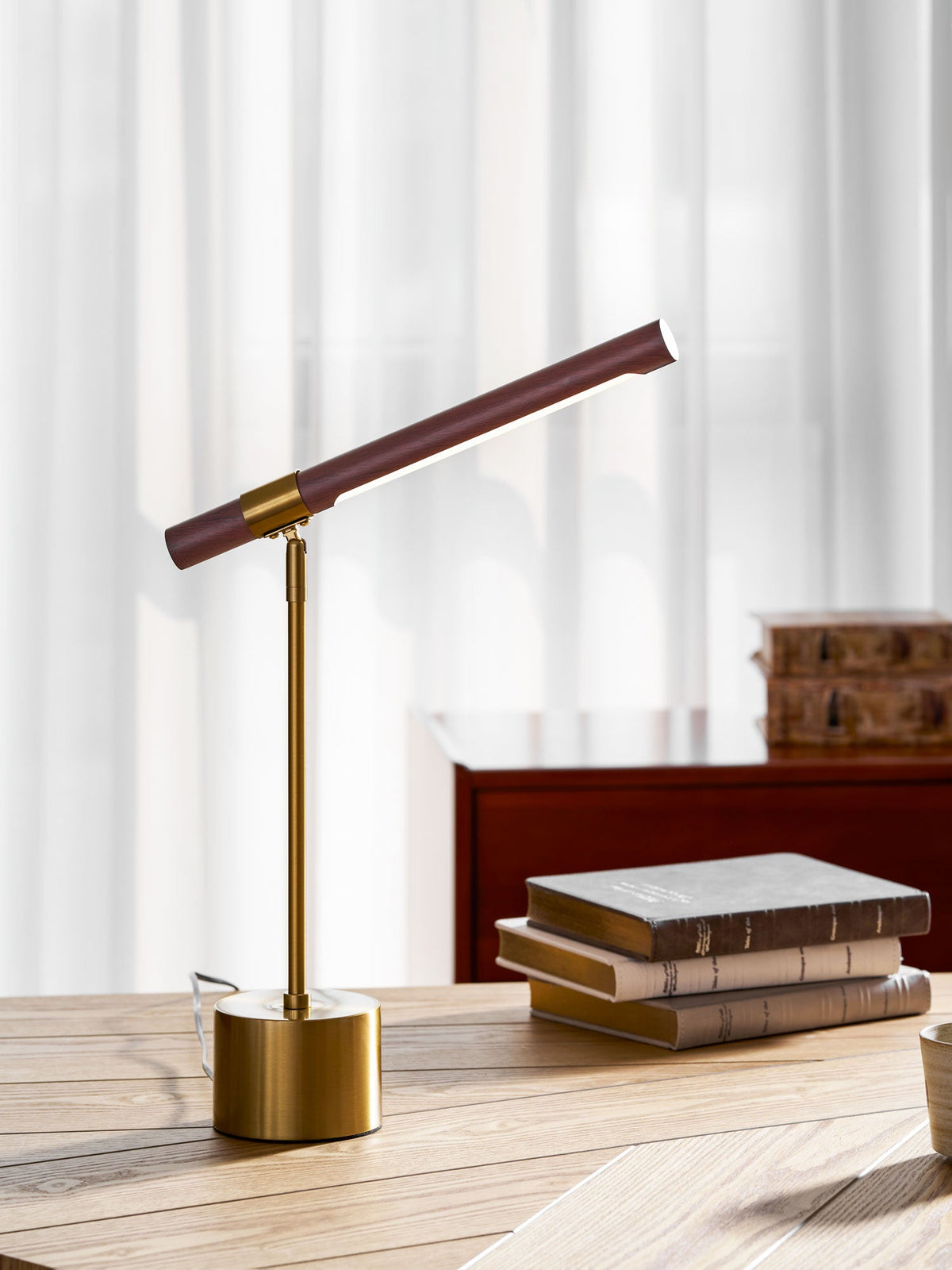 Clérachic table lamp on a herringbone desk, its bar raised towards a window of white curtains, with a stack of books to the right.