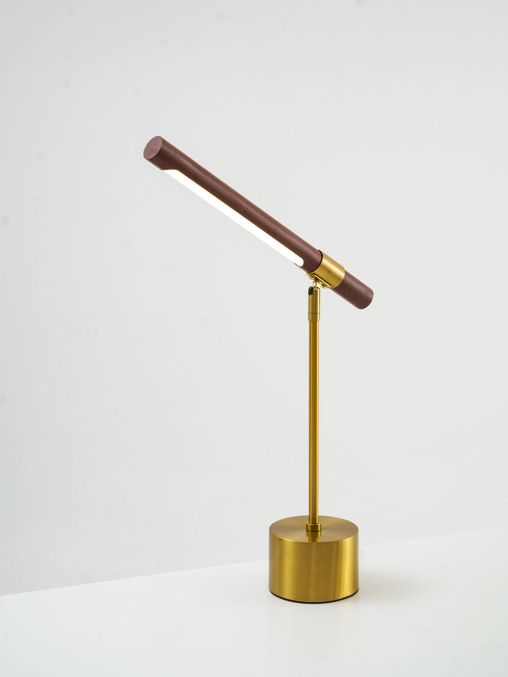 Clérachic lamp seen from a low side angle on a white surface, its walnut bar tilted steeply up and the brass base below.