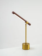 Clérachic lamp seen from a low side angle on a white surface, its walnut bar tilted steeply up and the brass base below.