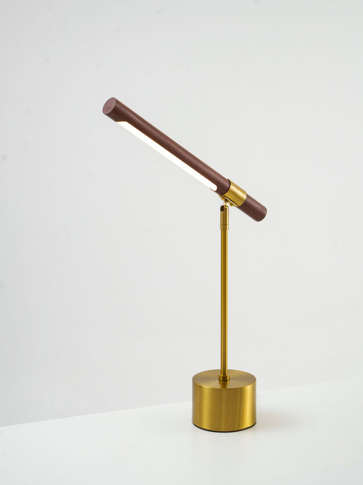 Clérachic lamp seen from a low side angle on a white surface, its walnut bar tilted steeply up and the brass base below.
