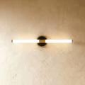 Clérachic slim LED tube wall light on a round black and brass plate, glowing warmly against a limewashed plaster wall.