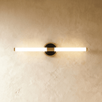 Clérachic slim LED tube wall light on a round black and brass plate, glowing warmly against a limewashed plaster wall.