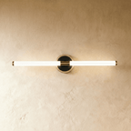 Clérachic longer LED tube wall light on a round black plate with a pale nickel-toned rim, lit against a plaster wall.