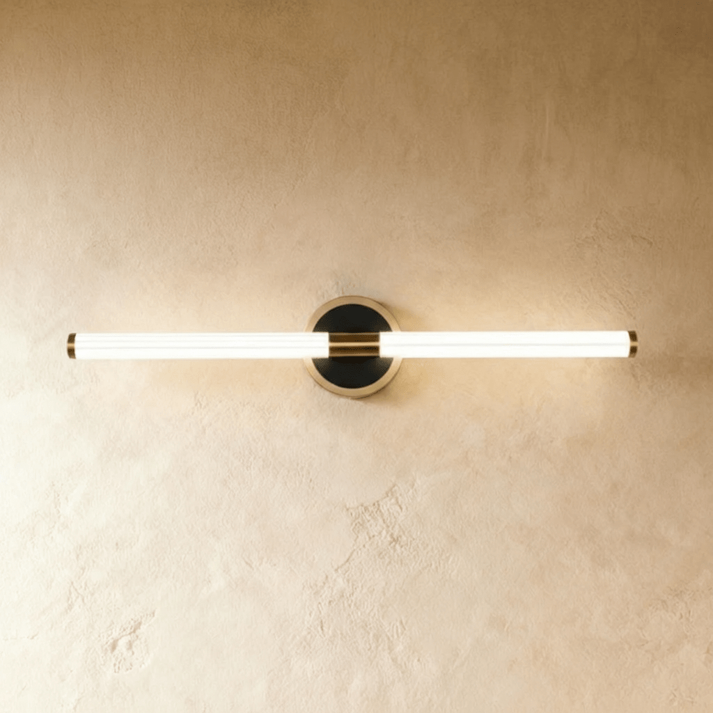 Clérachic longer LED tube wall light on a round black plate with a pale nickel-toned rim, lit against a plaster wall.
