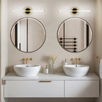 Pair of Clérachic LED tube wall lights on round brass plates above two round mirrors and white basins in a bathroom.