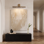A Clérachic picture light in bronze hung above a textured cream artwork and a black stone console in a pale hallway.