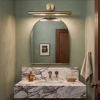 Bronze bar light mounted over an arched mirror in a green-walled bathroom with a marbled stone vanity, from Clérachic.