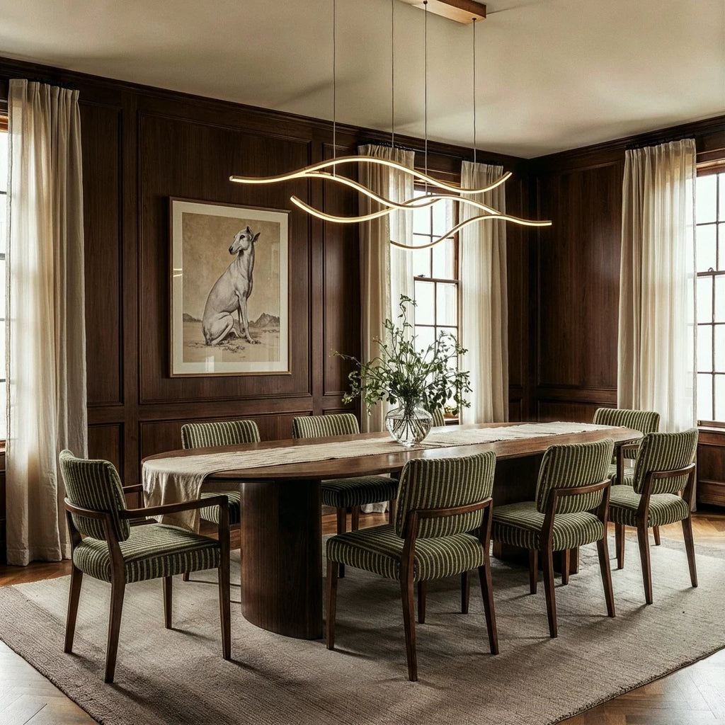 Clérachic gold wavy LED pendant glowing above a long walnut dining table in a panelled room with striped green chairs.