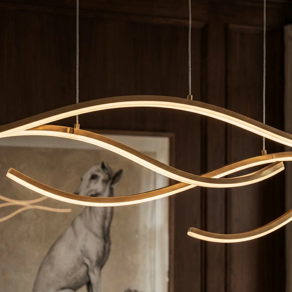 Close view of a Clérachic gold wavy LED pendant, its silicone diffuser glowing along slim aluminium arcs on fine cables.