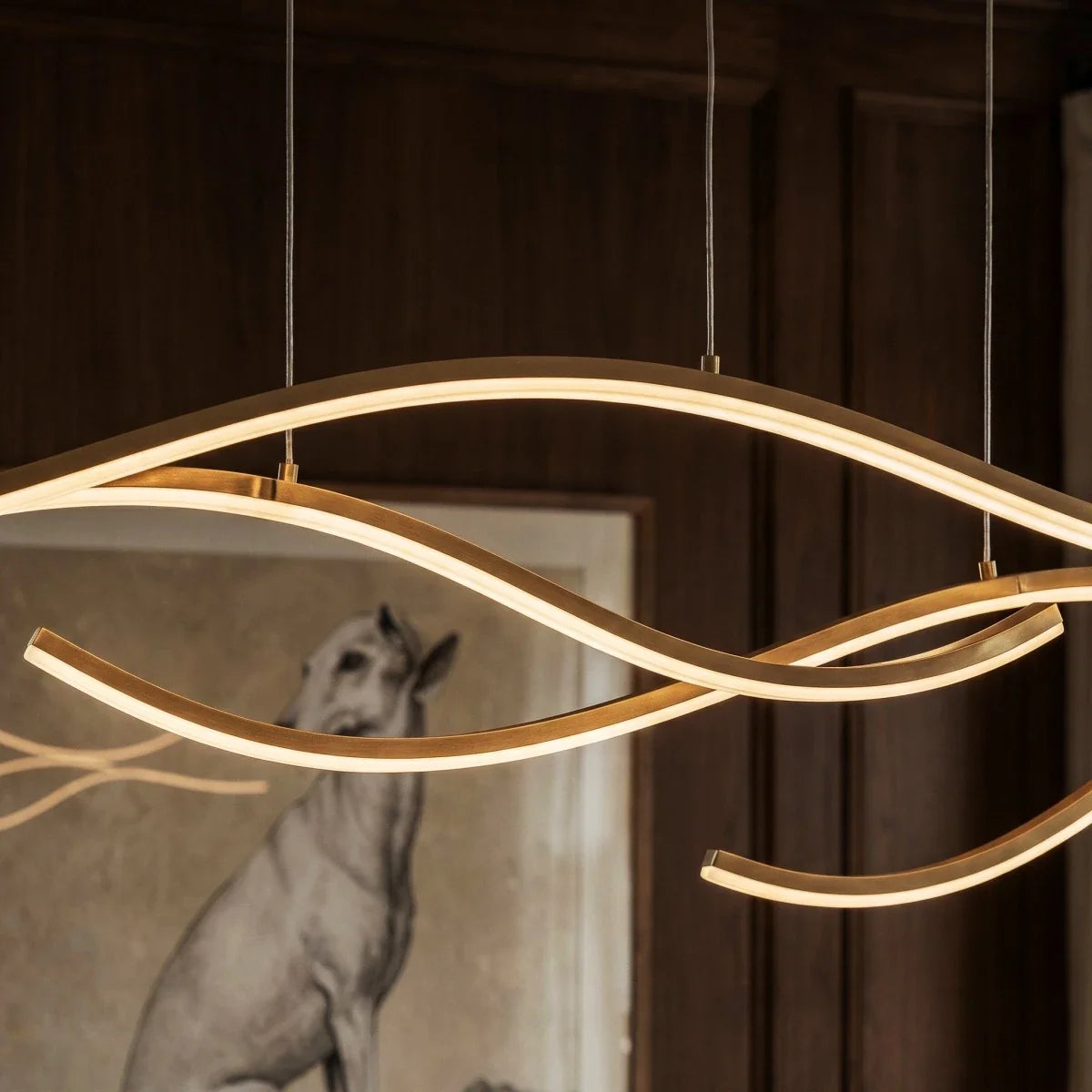 Close view of a Clérachic gold wavy LED pendant, its silicone diffuser glowing along slim aluminium arcs on fine cables.