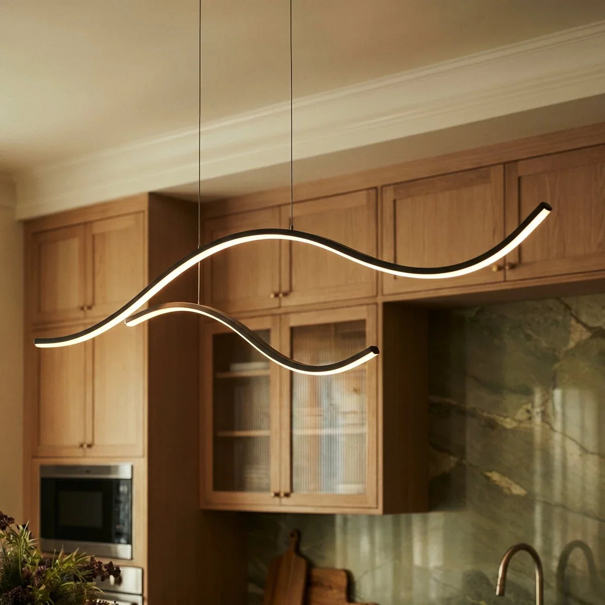 Close view of a Clérachic black wavy LED pendant, two glowing arcs on thin black cables in front of oak kitchen cabinets.