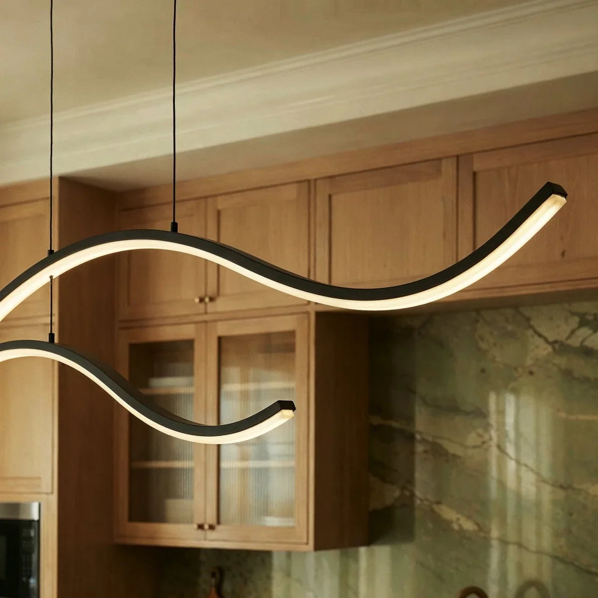 Clérachic black wavy LED pendant arcs in close-up, with the diffuser lit along the lower edge above oak glazed cabinets.