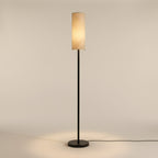 Clérachic slim floor lamp lit and seen straight on, its tall oatmeal linen shade glowing warm above a black iron pole and round disc base