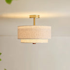 Lit Clérachic gold and linen flush-mount light, its brass stem glowing on a cream ceiling above a warm, softly lit wall.