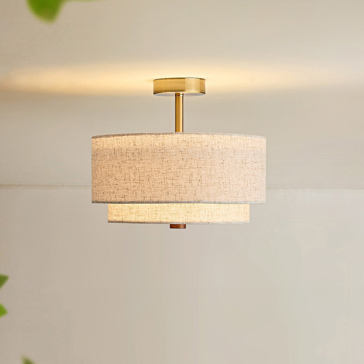Lit Clérachic gold and linen flush-mount light, its brass stem glowing on a cream ceiling above a warm, softly lit wall.