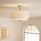 Clérachic gold and linen flush-mount light lit warm, with a brass canopy and stem, in front of an arched window and framed art.