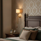 A lit beige-shaded Clérachic wall light hangs on damask wallpaper above a bedside table and a dark wooden bed with green and taupe cushions.